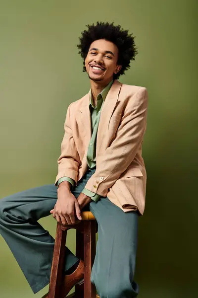 A stylish young African American man with curly hair sits atop a wooden stool, donning trendy attire on a green background. — Stock Photo