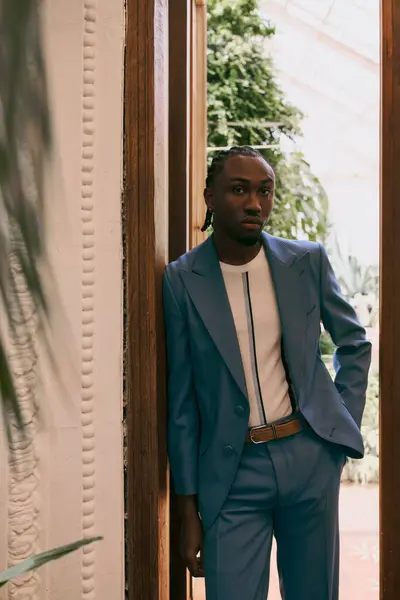 Beau Afro-Américain dans un costume élégant se penche contre une porte, exsudant sophistication dans un jardin vert vif. — Photo de stock