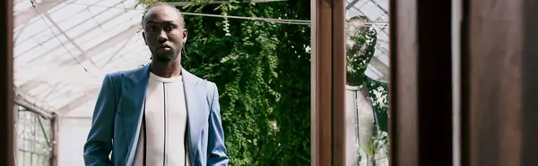 A handsome African American man in sophisticated dapper style stands in front of a window, framed by a vivid green garden. — Stock Photo