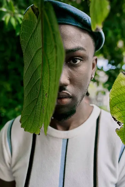 Hombre guapo con sombrero y tirantes está de pie con confianza junto a un árbol exuberante. - foto de stock