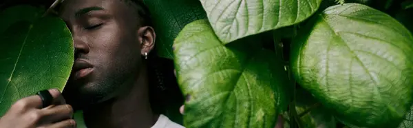 A handsome African American man in dapper style hides behind a large green leaf in a vivid garden. — Stock Photo