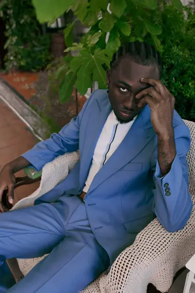 A handsome African American man in a blue suit sits on a chair in a vibrant green garden. — Stock Photo