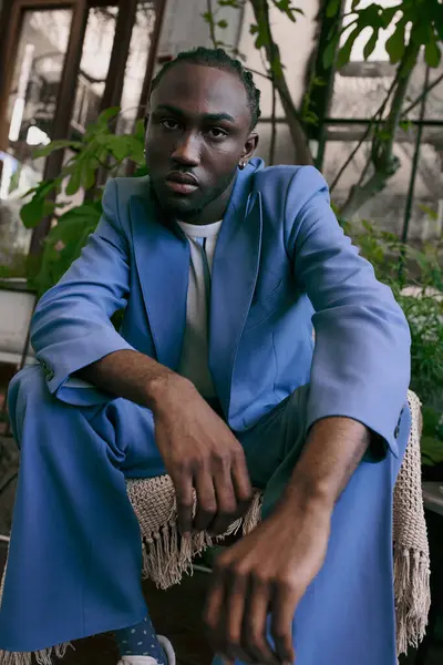 Un sofisticato uomo afroamericano in abito blu seduto con grazia su una sedia in un vibrante giardino verde. — Stock Photo