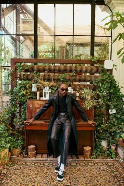 Стильний афроамериканський чоловік граціозно сидить на піаніно в кімнаті, наповненій яскравими рослинами. — стокове фото