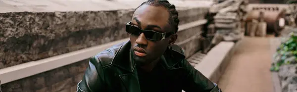 Schöner afroamerikanischer Mann in schwarzer Lederjacke und Sonnenbrille posiert in einem grünen Garten. — Stockfoto