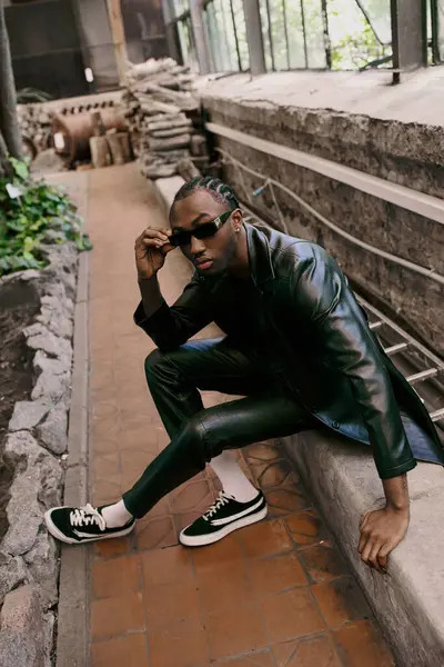 Handsome African American man in black leather jacket and sunglasses sitting on ledge. — Stock Photo