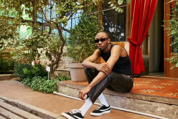 Hombre afroamericano guapo con estilo elegante sentado con las piernas cruzadas en un paso en un jardín verde vibrante. - foto de stock