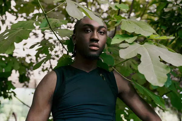 An African American man with sophisticated style stands gracefully in front of a lush tree in a vivid green garden. — Stock Photo