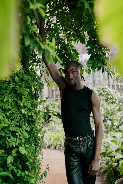 Sophisticated African American man in black shirt and leather pants standing under lush tree. — Stock Photo