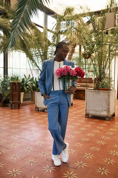 Hombre afroamericano guapo en traje azul sosteniendo flores en un jardín verde vivo. - foto de stock