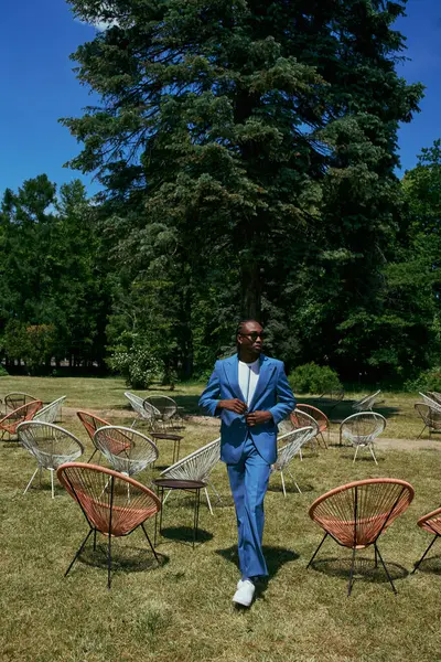 Bonito homem de terno azul sofisticado em pé em um campo de cadeiras em um jardim verde vívido. — Fotografia de Stock