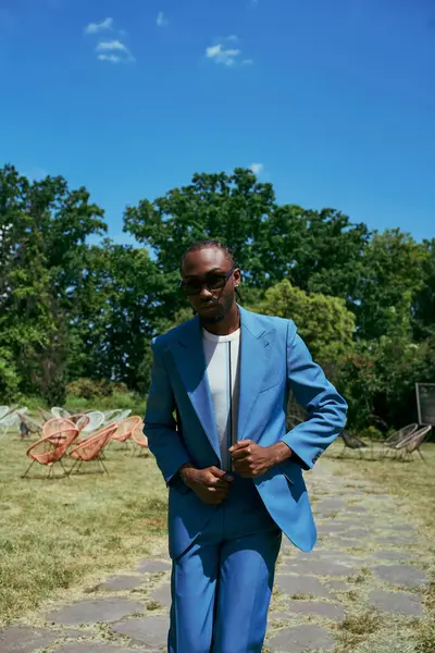 Handsome African American man in a blue suit and sunglasses poses in a vivid green garden. — Stock Photo