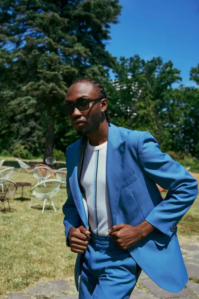 Un homme afro-américain sophistiqué pose dans un jardin vert vif en costume bleu et lunettes de soleil. — Photo de stock