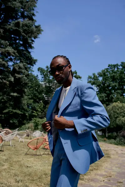 Beau Afro-Américain en lunettes de soleil et costume bleu posant dans un jardin vert vif. — Photo de stock
