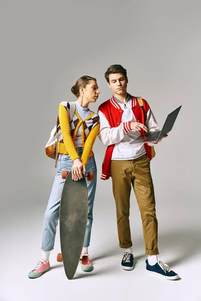 A young male and female students hold a skateboard and laptop, standing in a casual pose. — Stock Photo
