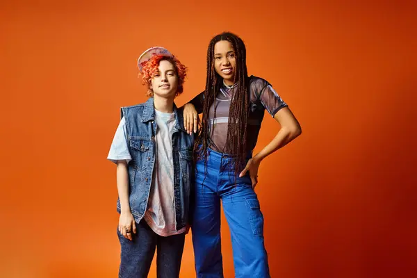 Two stylishly dressed young multicultural friends, including a nonbinary person, standing confidently next to each other in front of a bold orange backdrop. — Stock Photo