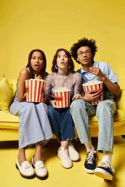 Drei Menschen unterschiedlichster Herkunft sitzen auf einer stylischen Couch, halten Popcornboxen in der Hand und genießen die Gesellschaft des jeweils anderen. — Stockfoto