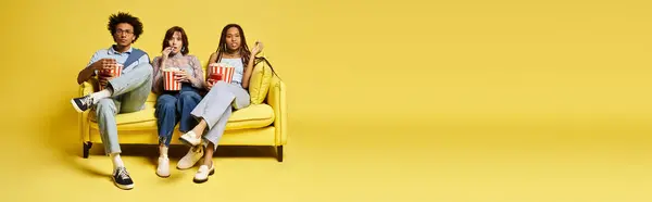 A group of young multicultural friends, including a nonbinary person, sit stylishly on top of a bright yellow chair in a studio setting. — Stock Photo