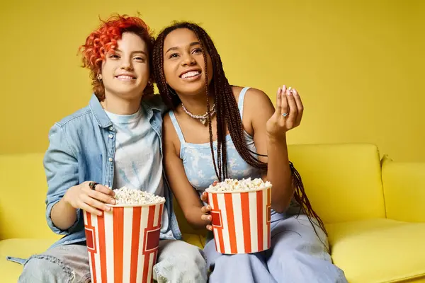 Zwei junge Leute in stylischer Kleidung sitzen auf einer Couch und halten Popcorn-Eimer in der Hand und genießen einen gemeinsamen Filmabend. — Stockfoto