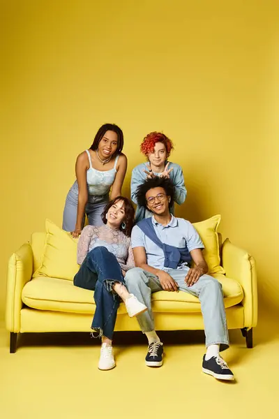 A group of young multicultural friends in stylish attire sitting together on a yellow couch in a studio setting. — Stock Photo