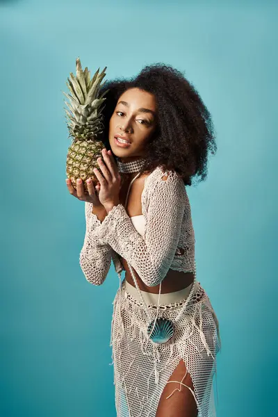 Young African American woman in trendy swimsuit holding pineapple on blue backdrop. — Foto stock