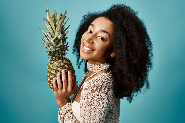 Young African American woman in swimsuit holding pineapple against blue backdrop. — Foto stock