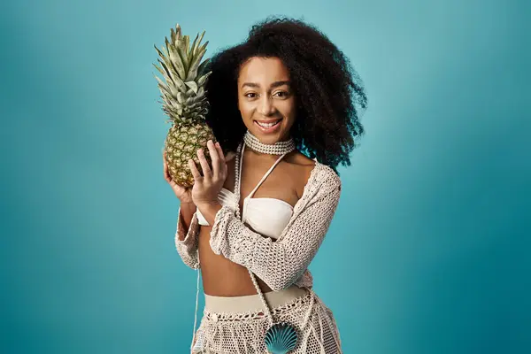 Attractive African American woman holding pineapple in trendy swimsuit against blue backdrop. — Foto stock