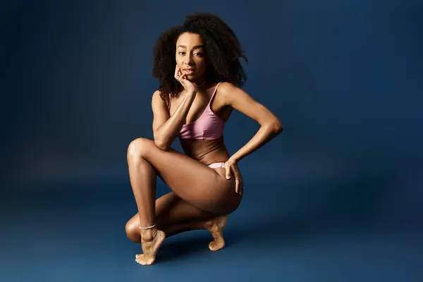Young African American woman crouching in chic pink swimwear on blue background. — Stock Photo