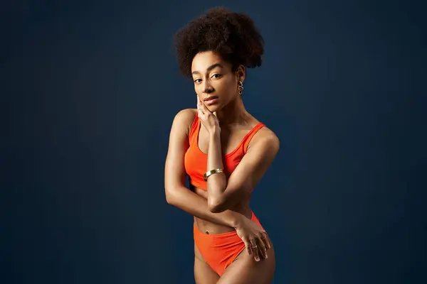 Stylish African American woman poses in orange bikini against blue backdrop. — Stock Photo