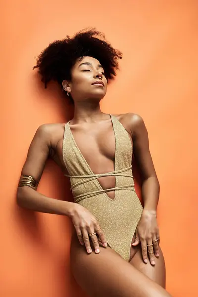 Femme afro-américaine élégante en maillot de bain en or frappe une pose sur fond orange vif. — Photo de stock