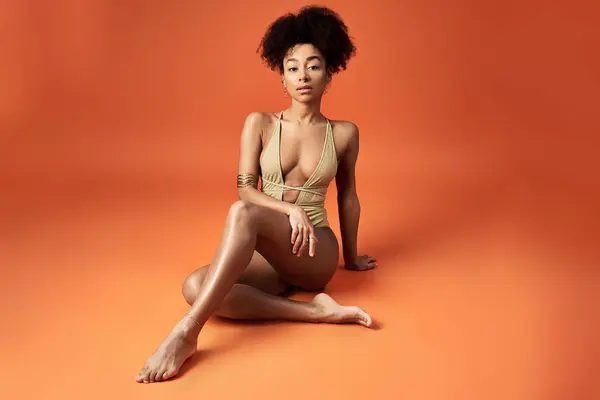 African American woman in trendy swimsuit, sitting elegantly on bright orange backdrop. — Stock Photo