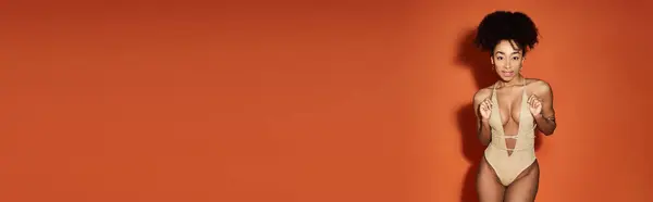 African American woman in trendy swimsuit striking a pose against bright orange backdrop. — Stock Photo