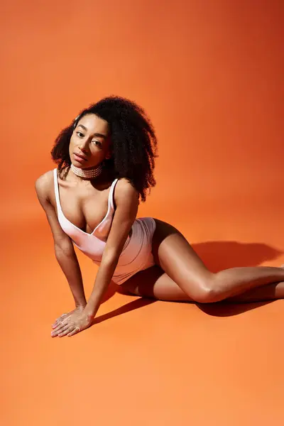 Stylish African American woman poses in white swimsuit on vibrant orange backdrop. — Stock Photo