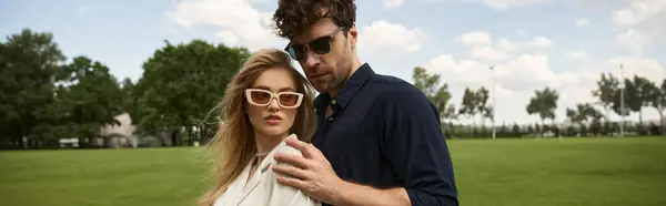 Um casal elegante em óculos de sol está em um campo exuberante, exalando elegância e confiança em seu traje na moda. — Fotografia de Stock