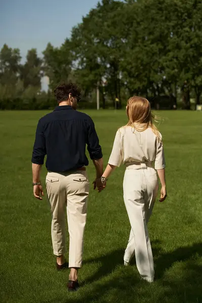 A young couple in elegant attire holding hands while walking through a lush park together. — стокове фото