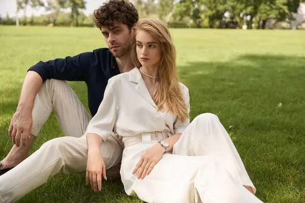 A stylish, young man and woman in elegant attire sit closely together on the lush green grass. — Stock Photo