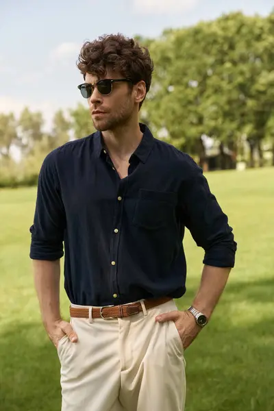 Um homem elegante vestido com uma camisa da marinha e calças bronzeadas de pé graciosamente em um campo verde. — Fotografia de Stock