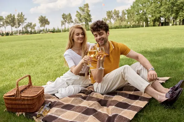 A sophisticated man and woman in stylish outfits sitting on a blanket near a picnic basket in a lush park. — стокове фото