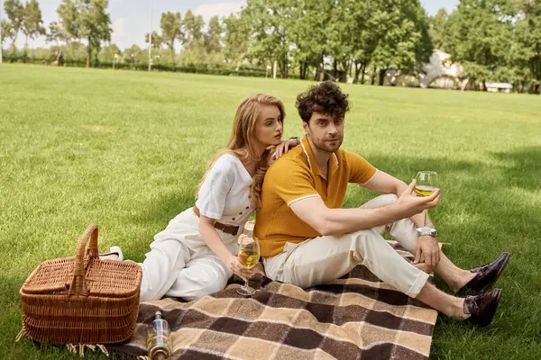 Um homem e uma mulher em roupas elegantes sentam-se em um cobertor em um parque, desfrutando de uma garrafa de vinho juntos sob o sol quente.. — Fotografia de Stock