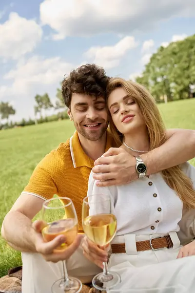 Una bella coppia giovane in eleganti brindisi di abbigliamento con bicchieri di vino in un parco lussureggiante, incarnando lusso e raffinatezza. — Foto stock