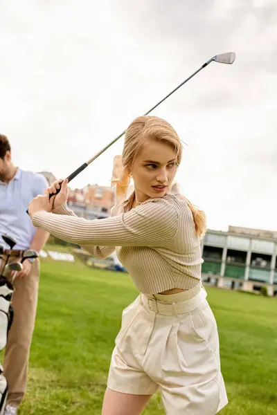 A woman in elegant attire swings a golf club in front of a man on a green field, embodying an upper-class pastime. — стокове фото
