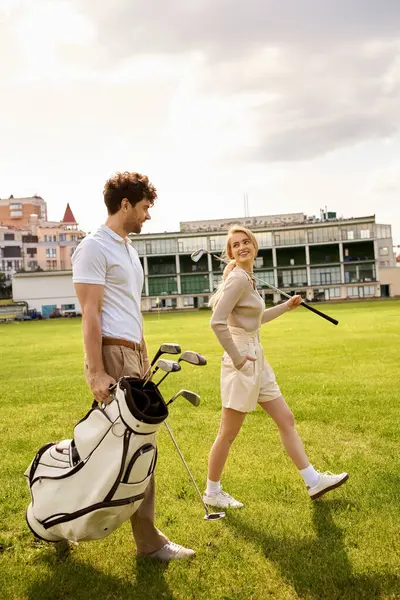 A man and woman in elegant attire walking casually on a grassy field with golf clubs in hand. — стокове фото