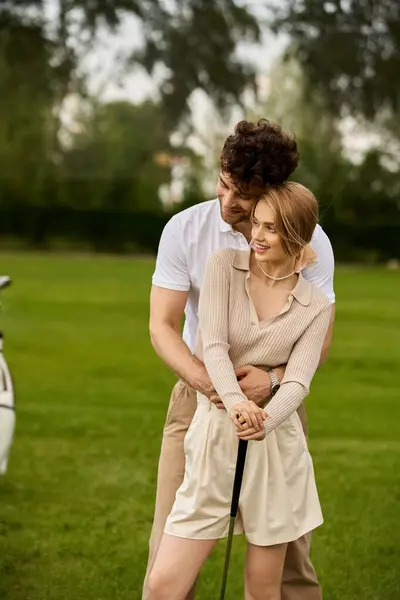 Un hombre y una mujer en traje elegante abrazan tiernamente en un campo de golf verde exuberante, tomando el sol en la tranquilidad de su momento juntos. - foto de stock