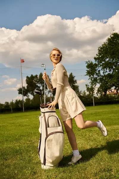 Uma mulher de traje elegante fica em um campo gramado com um saco de golfe, pronto para jogar em um dia ensolarado no clube de golfe. — Fotografia de Stock