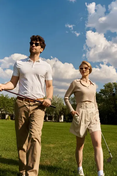 Ein stilvoller Mann und eine stilvolle Frau schlendern über ein lebhaftes Feld und halten Golfschläger unter dem klaren Himmel eines gemütlichen Nachmittags.. — Stockfoto