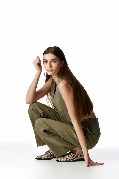 A beautiful woman with long dark hair kneels gracefully in a stylish green jumpsuit against a gray backdrop. — Stock Photo