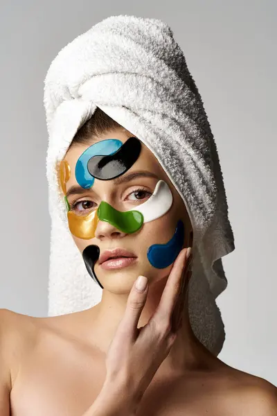 Una mujer joven posa con una toalla envuelta alrededor de su cabeza, luciendo manchas coloridas en los ojos. - foto de stock