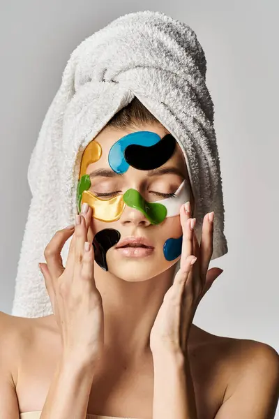 Una joven serena y elegante con parches en la cara, mostrando una rutina de belleza con toallas envueltas alrededor de su cabeza. - foto de stock