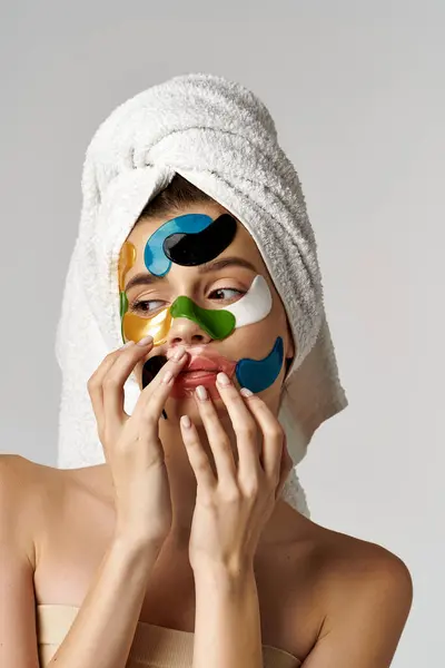 Una joven con una toalla envuelta alrededor de su cabeza mostrando parches para los ojos adornados con maquillaje y productos para el cuidado de la piel. - foto de stock