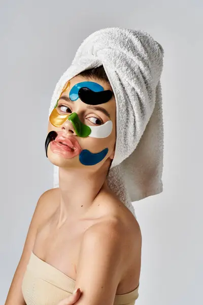 Una hermosa joven posa con una toalla envuelta alrededor de su cabeza y parches intrincados en los ojos. - foto de stock